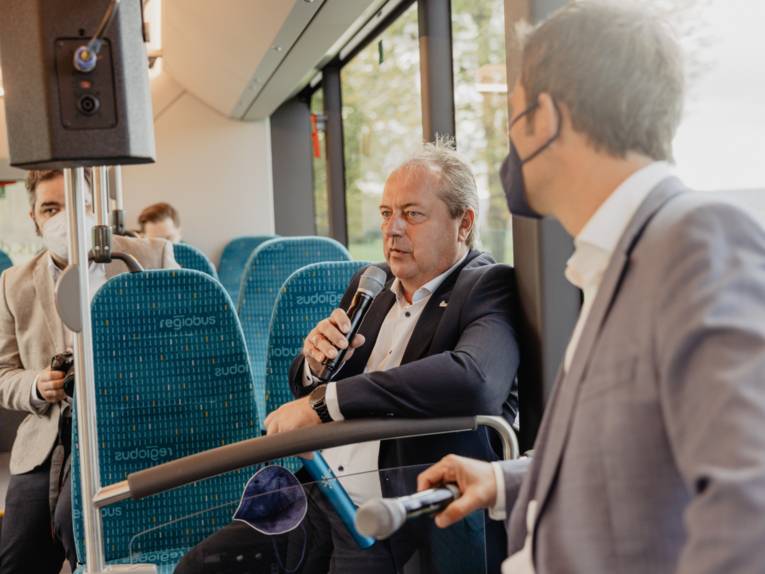 Passagiere in einem Bus, einer hält ein Mikrofon in der Hand und spricht hinein